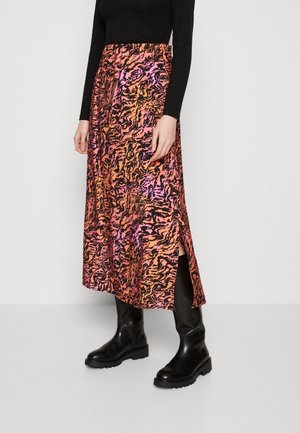 Femme portant une jupe mi-longue orange et rose à motifs avec une fente latérale, un top noir à manches longues et des bottes noires montant jusqu'au genou.