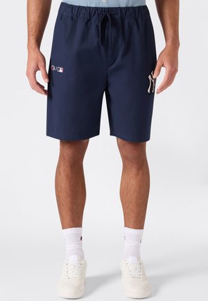 Mann trägt marineblaue Sportshorts mit MLB- und New York Yankees-Logos, weiße Socken und weiße Sneakers und steht vor weißem Hintergrund.