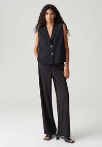 Schwarze ärmellose Weste mit Reverskragen, Ein-Knopf-Verschluss und Fronttaschen, kombiniert mit hoch taillierten schwarzen Hosen und offenen Mules.