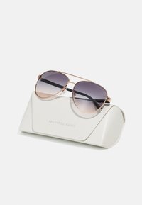 Sonnenbrille mit verlaufenden Gläsern, roségoldenem Metallrahmen und schwarzen Kunststoffbügeln, die auf einem strukturierten weißen Etui mit geprägtem Logo liegt.