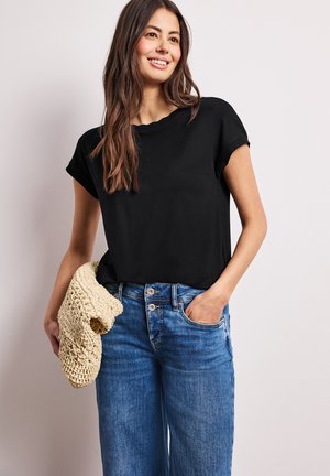 Ung kvinde smilende iført en sort t-shirt med korte ærmer og blå jeans, holder et beige strikket stykke i den ene hånd.