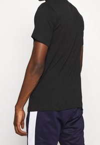 Man som bär en svart kortärmad skjorta och mörkblå byxor med vita sidostreck, stående mot en enkel vit bakgrund.