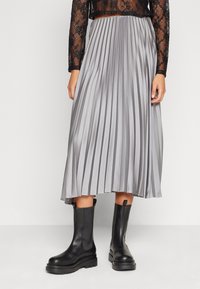 Jupe midi plissée en tissu argenté avec une texture lisse, associée à des bottines noires à semelle robuste et à design droit.