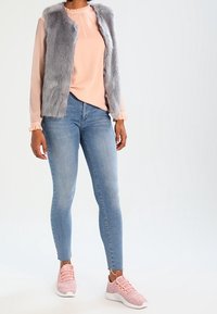 Haut rose clair à manches longues avec manches à volants et gilet en fausse fourrure grise, associé à un jean bleu slim et des baskets de sport roses.