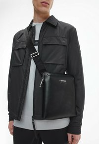 Borsa messenger nera in pelle, forma rettangolare, con zip e logo Calvin Klein. Texture liscia, tracolla staccabile per una maggiore versatilità.