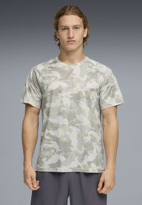 Νέος με σγουρά μαλλιά που φοράει κοντομάνικο γκρι πουκάμισο με καμουφλάζ και σκούρο γκρι παντελόνι, στέκεται μπροστά σε γκρίζο φόντο.