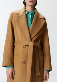 Kaput od vune u boji kamila sa širokim reverima, pojasom u struku i dva dugmeta. Ispod je naglašen tirkiznom košuljom. Vidljive bočne džepove.