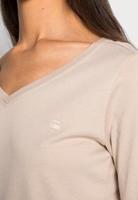 Beige V-ringad skjorta i mjukt tyg med en subtil logotyp på bröstet. Har en avslappnad passform och ren sömnad längs halsringningen.