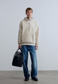 Μπεζ φούτερ με κουκούλα και μπροστινή τσέπη, μπλε τζιν τζιν και μαύρα παπούτσια. Κρατά μια μαύρη ταξιδιωτική τσάντα, στέκεται μπροστά σε ανοιχτό γκρι φόντο.