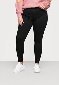 Personne portant un pantalon slim noir, des baskets blanches et un pull rose, debout devant un fond clair uni.