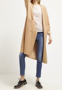 Cardigan largo beige con apertura frontal, dos grandes bolsillos, combinado con una blusa ligera y jeans ajustados azules. Zapatos negros con cordones completan el look.