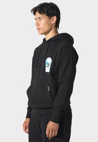 Joven con sudadera negra con gráfico de coche en el pecho y pantalones negros a juego de pie de lado contra un fondo sencillo.