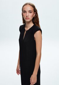 Vestido negro sin mangas con escote en forma de llave, detalles plisados y silueta ajustada, hecho de una tela suave con un brillo sutil.