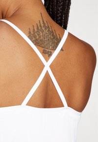 Weißes Tanktop mit dünnen überkreuzten Trägern, das eine glatte Textur und ein schlichtes Design aufweist. Ein Tattoo ist auf dem oberen Rücken sichtbar.