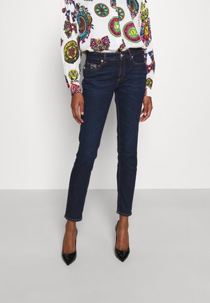 Kvinde iført mørke skinny jeans, sorte spidse højhælede sko og en hvid langærmet bluse med farverige cirkelmønstre.