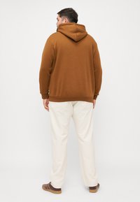 Brązowy hoodie z kieszenią kangurzą i kapturem ze sznurkiem, zestawiony z jasnymi kremowymi spodniami. Widoczne detale szwów na spodniach.
