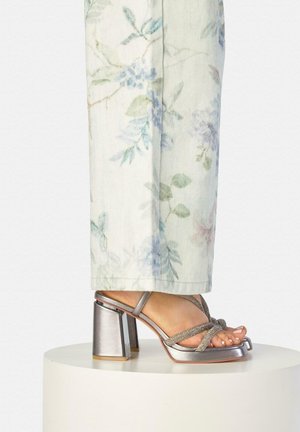 Pie calzado con sandalia plateada de tacón cuadrado con tiras brillantes, combinada con pantalones de color claro con un sutil estampado floral.