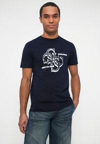 Hombre vestido con camiseta gráfica azul marino y jeans azules, de pie con una mano en el bolsillo frente a un fondo blanco.