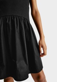 Černé šaty s přiléhavým žebrovaným topem a rozšířenou sukní. Vyrobeny z měkké, strukturované tkaniny s jemným leskem, délka ke kolenům na modelce.