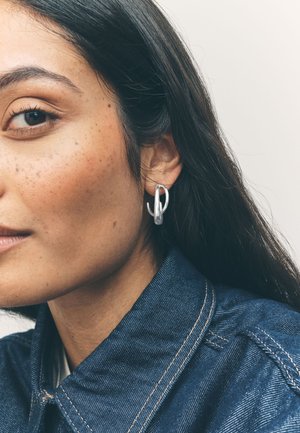 Close-up of a person with long dark hair wearing a silver abstract-shaped earring and a blue denim jacket against a plain background.