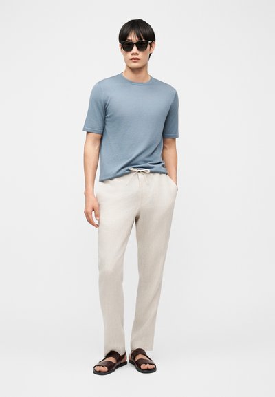 Άνδρας που φορεί γαλάζιο κοντομάνικο πουκάμισο, μπεζ παντελόνι με κορδόνι, σκούρα σανδάλια και μαύρα γυαλιά ηλίου, στέκεται μπροστά σε απλό φόντο.