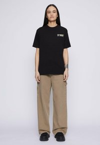 Pikkade tumejuustega inimene seisab seljas must trükitud T-särk, beežid laiad püksid, mustad kingad ja tätoveering vasakul alumise käe peal.