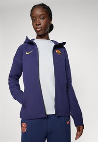 Lila Zip-Hoodie mit erhöhtem Kragen, gelbem Nike-Logo und Barcelona-Wappen. Begleitet von navyblauen Hosen mit Logo-Details.