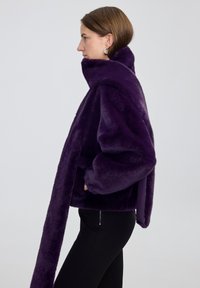 Donna di profilo che indossa una giacca in pelliccia sintetica di un profondo colore viola con un collo oversize e una lunga sciarpa abbinata, abbinata a pantaloni neri.