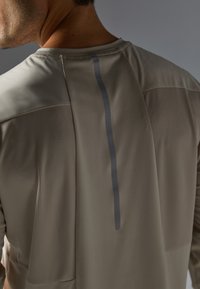 Chemise à manches courtes de couleur gris clair avec un motif texturé. Caractérisée par une bande réfléchissante verticale dans le dos et un col rond.