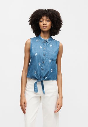 Žena s kučeravými vlasmi oblečená v modrom tielkovom top s gombíkmi a bielym kvetinovým vzorom, zviazaným vpredu, v kombinácii s bielymi nohavicami, stojaca pred bielym pozadím.