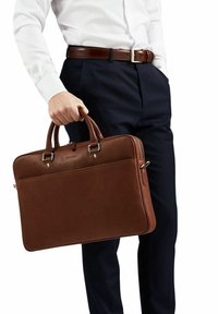 Brun læder arbejdstaske med dobbelt top håndtag, sølvhardware, en glat tekstur og lynlås lukning, holdt af en person i formelt tøj.
