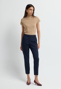 Kvinna som står, klädd i en beige kortärmad stickad topp, marinblå skräddarsydda byxor och bordeauxfärgade låga klackskor mot en ljus enfärgad bakgrund.