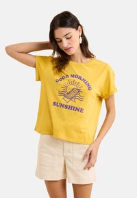 T-shirt en coton jaune à manches courtes et design court, avec un texte violet et un graphique représentant un soleil et des vagues.