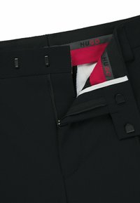 Schwarze Hosen aus strukturiertem Stoff, mit einem Frontreißverschluss, Hakenverschluss und einem kontrastierenden roten und weißen Innenfutter mit Markenlogo.