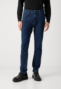 Temno modri traper hlače s ravnim krojem, petimi žepi in klasičnim gumbom ter zadrgo, v kombinaciji s črnimi usnjenimi čevlji.