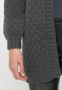 Mørk grå strikket cardigan med et teksturert mønster og ribbede mansjetter og kanter. Materialet ser mykt og behagelig ut.