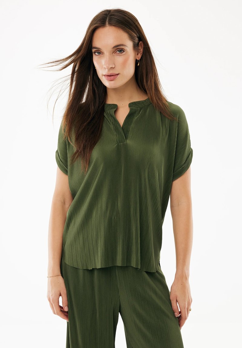 Frau mit langen braunen Haaren, die ein lockeres, strukturiertes olivgrünes Kurzarmoberteil und passende Hosen trägt, vor einem weißen Hintergrund stehend.