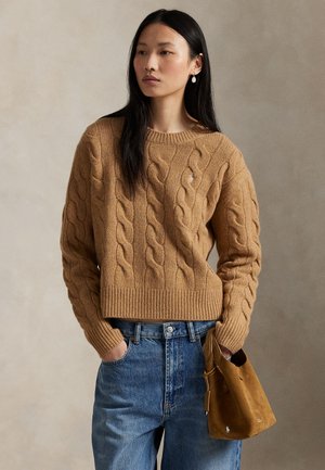 Kobieta o długich czarnych włosach, ubrana w kamelowy sweter warkoczowy i niebieskie dżinsy, trzymająca brązową zamszową torebkę na jednolitym tle.