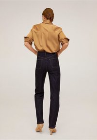 Dark blue denim high-waisted jeans with back pockets, paired with a tan satin blouse, visible cuffs rolled up. Model wearing nude heels.