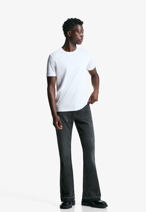 Weißes Baumwoll-T-Shirt kombiniert mit dunkelgrauen Schlagjeans und schwarzen Schuhen. Das Outfit besticht durch ein einfaches Design und eine lockere Passform.