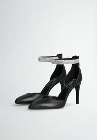 Scarpe nere in pelle con tacco alto, punta appuntita, design con ritagli e cinturino alla caviglia adornato con strass. Texture liscia e silhouette elegante.