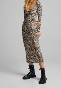 Žena v midi šatech s dlouhým rukávem, řasených, s hnědými, černými a modrými vírovými vzory, doplněná černými šněrovacími vojenskými botami.