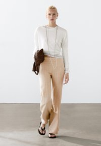 Lichtgrijze lange mouw top, beige gestreepte wijde broek, zwarte sandalen, houdt een bruine geweven tas vast, met een eenvoudige ketting als accent.