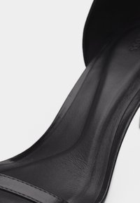 Gros plan de la semelle intérieure en cuir noir et de la bride d'une chaussure à talon haut sur un fond clair uni.