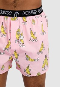 Pink boxer shorts featuring a yellow banana cartoon pattern with smiling faces. Black elastic waistband with "LOUSY LIVIN" text. Soft cotton material.