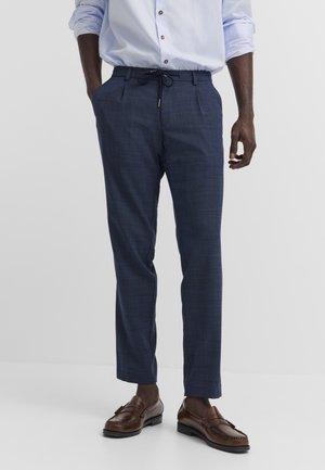 Mann trägt marineblaue, maßgeschneiderte Hose mit Kordelzugbund, ein hellblau gestreiftes Hemd mit Knöpfen und braune Leder-Loafer.