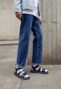 Jeans de mezclilla anchos de color azul, chaqueta azul claro, sandalias en blanco y negro con tiras, y una blusa en capas blanca y gris. Fondo de concreto.