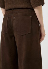 Pantaloni a gamba larga in camoscio marrone con due tasche posteriori, caratterizzati da dettagli in metallo argentato e cuciture visibili lungo le cuciture.
