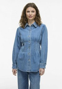 Cămașă din denim cu închidere cu nasturi, de culoare albastru deschis, mâneci lungi, talie ajustată și pliseuri laterale. Dispune de un guler clasic și un tiv rotunjit.