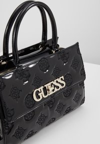 Sac à main en cuir verni noir avec motif de logo en relief, quincaillerie dorée, forme structurée et double poignées arrondies.